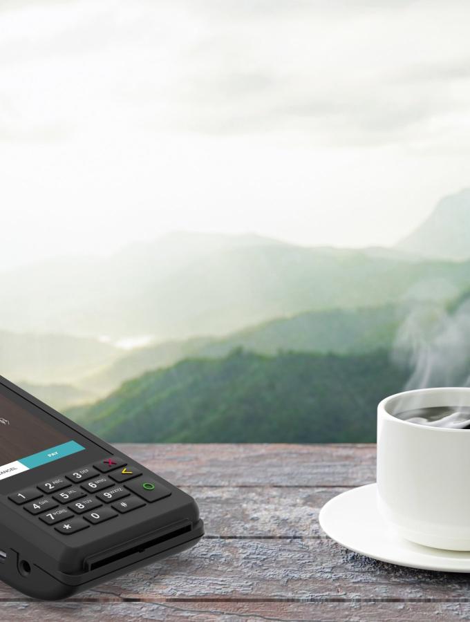 V210 payment device on an outdoor cafe table, coffe pouring into a cup, mountains in the background